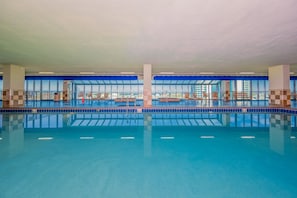 A large indoor swimming pool with clear blue water, surrounded by blue-tiled walls and windows offering views of buildings outside. - Phoenix Ix 1002 2 Bedroom Condo (Orange Beach)