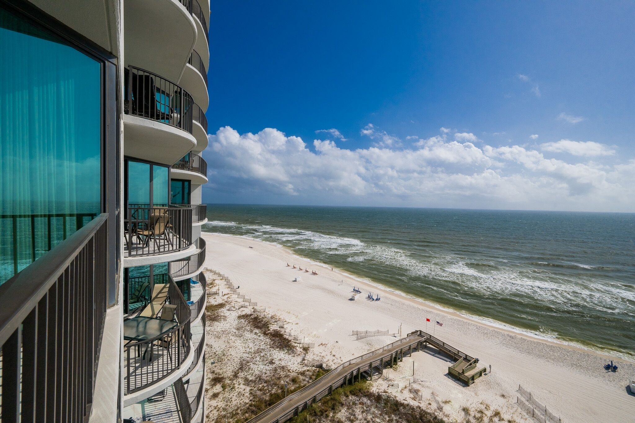 A beachfront hotel with multiple balconies overlooking the ocean, a sandy beach, and a wooden boardwalk leading to the water.