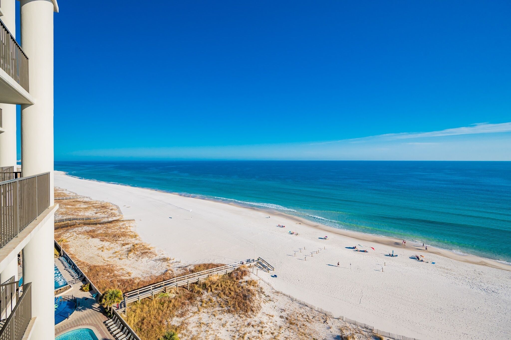 En strand med krystalklart hav, sandet bredde og en bygning med balkon med udsigt over stranden.
