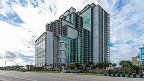 A modern multi-story building with a glass facade, a prominent entrance with a curved arch, and a parking lot in front. - The Oasis 1003 Formerly Phoenix West Ii 1003 3 Bedroom Condo (Orange Beach)