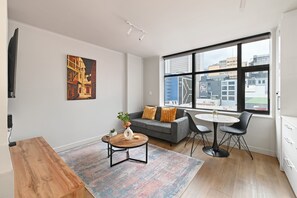 A modern living room with a grey sofa, a coffee table, and a dining area with chairs. There is a large window with a city view. - Stylish Roof Top Pool Studio Apartment The Tokyo (Cape Town)