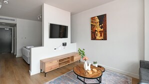 A modern living room with a flat-screen TV mounted on a white wall, a wooden bench with storage, a round coffee table with decorative items, and a colorful rug. - Stylish Roof Top Pool Studio Apartment The Tokyo (Cape Town)
