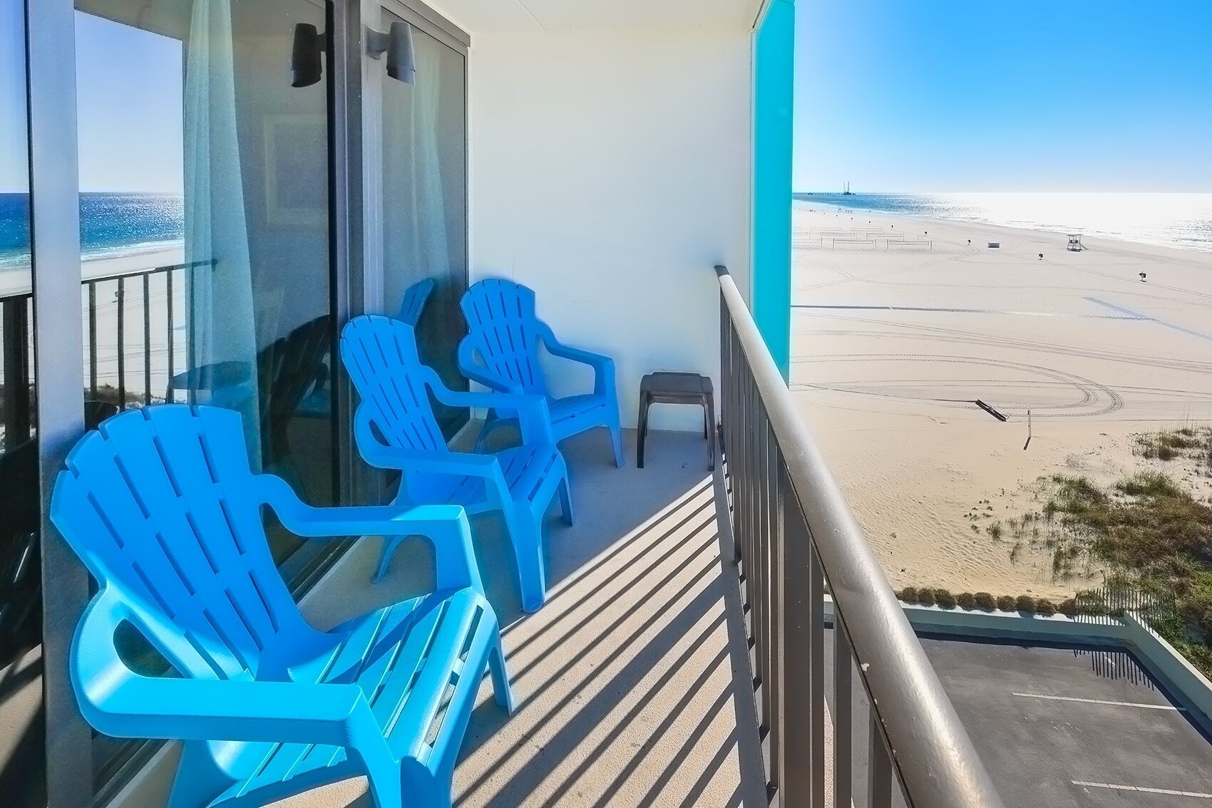 Un balcón con sillas azules, un taburete pequeño y vista a una playa con arena y un muelle a lo lejos.