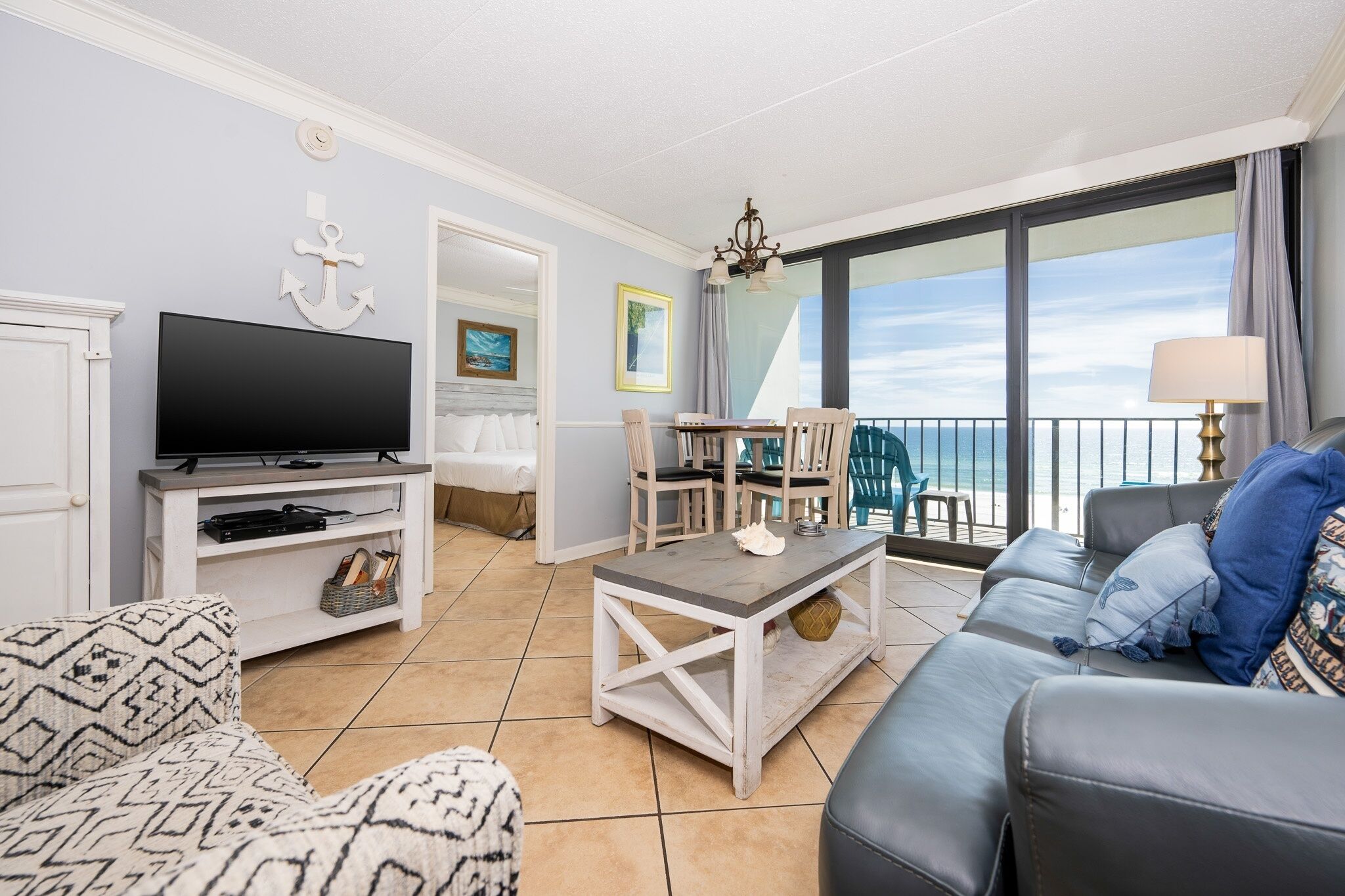 A living room with a television, a dining area with chairs, a sofa, and a balcony with a view of the sea.