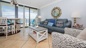 A living room with a balcony, a sofa, a coffee table, and a dining area with chairs. - Island Winds East 503 2 Bedroom Condo (Gulf Shores)