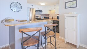 A kitchen with a bar area, white cabinets, a clock, and a framed picture on the wall. - Island Winds East 503 2 Bedroom Condo (Gulf Shores)