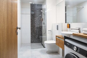 A modern bathroom with a glass shower enclosure, a toilet, a sink, and a washing machine. - My Second Home - Soukenická 36 Residence (Hlavní město Praha)