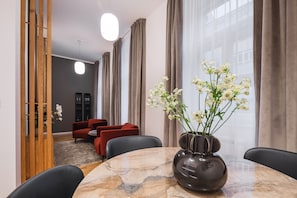 A modern hotel room with a marble dining table, red sofas, a vase of flowers, and a view of the city through large windows. - My Second Home - Soukenická 36 Residence (Hlavní město Praha)