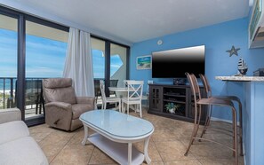 A balcony with a view of the beach, a flat-screen TV, and a seating area with a glass table and chairs. - Island Winds East 204 1 Bedroom Condo (Gulf Shores)