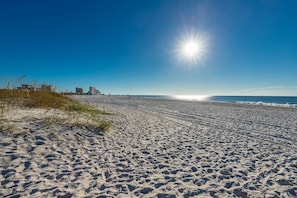 A wide sandy beach with footprints leading to the water, a clear sky, and buildings in the background. - Island Winds East 204 1 Bedroom Condo (Gulf Shores)