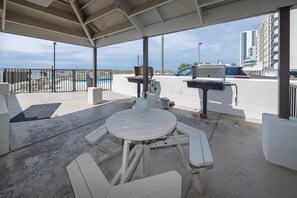 A covered outdoor area with a picnic table and chairs, a barbecue grill, and a view of a pool and buildings in the background. - Island Winds East 604 1 Bedroom Condo (Gulf Shores)
