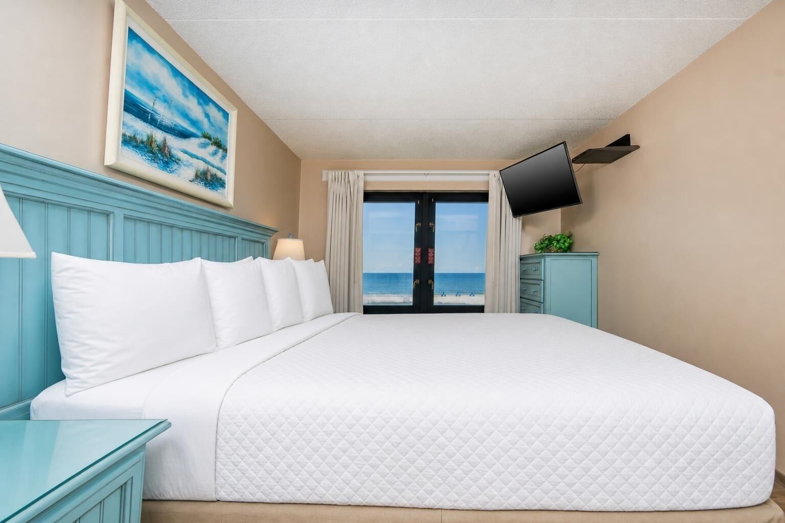 A hotel room with a large bed, a television mounted on the wall, and a view of the beach through a door.