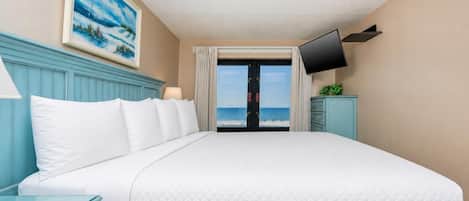 A hotel room with a large bed, a television mounted on the wall, and a view of the beach through a door.
