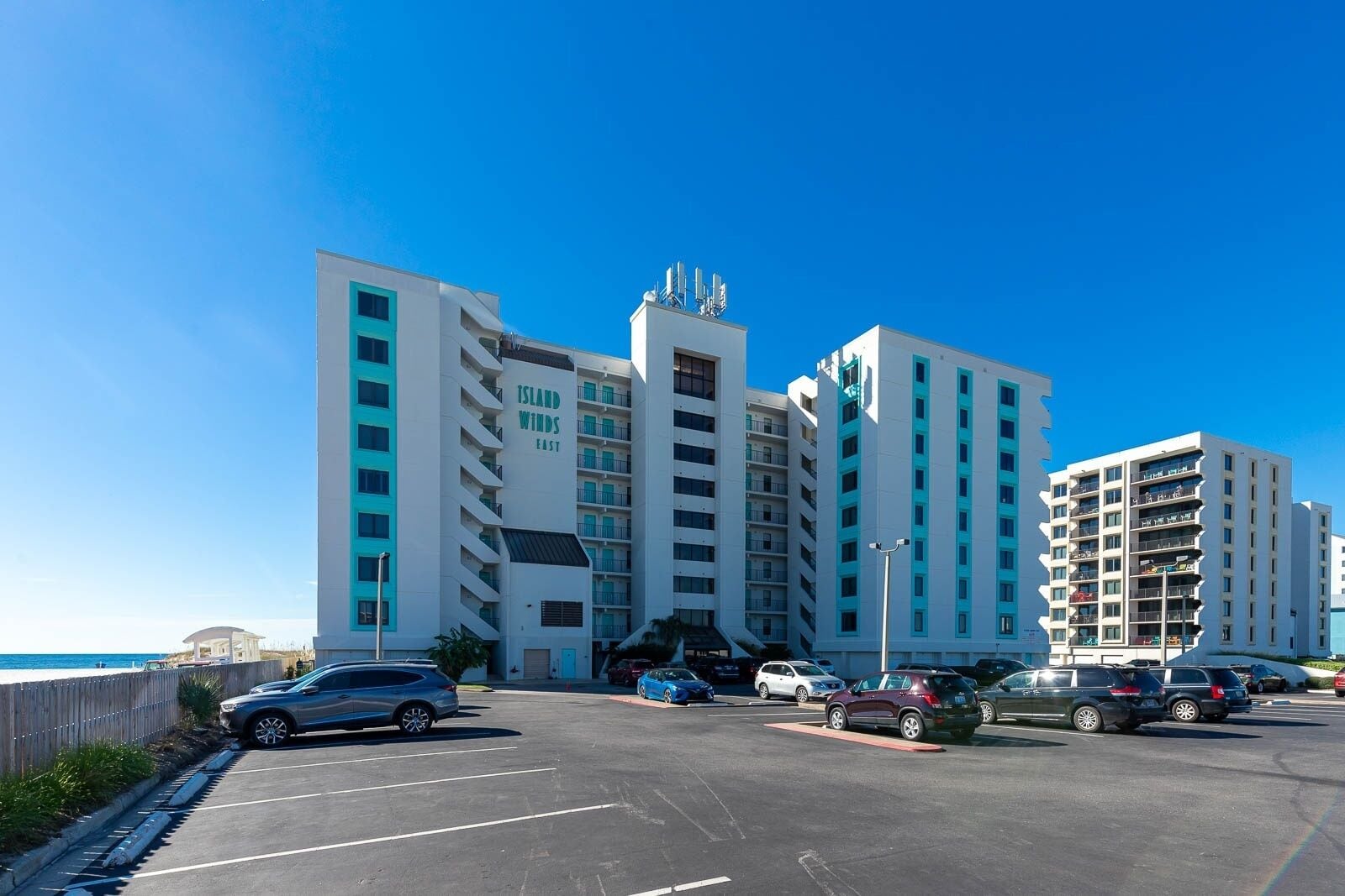 A multi-story hotel with a prominent 'Island Winds East' sign, a parking lot with several cars, and a clear blue sky.