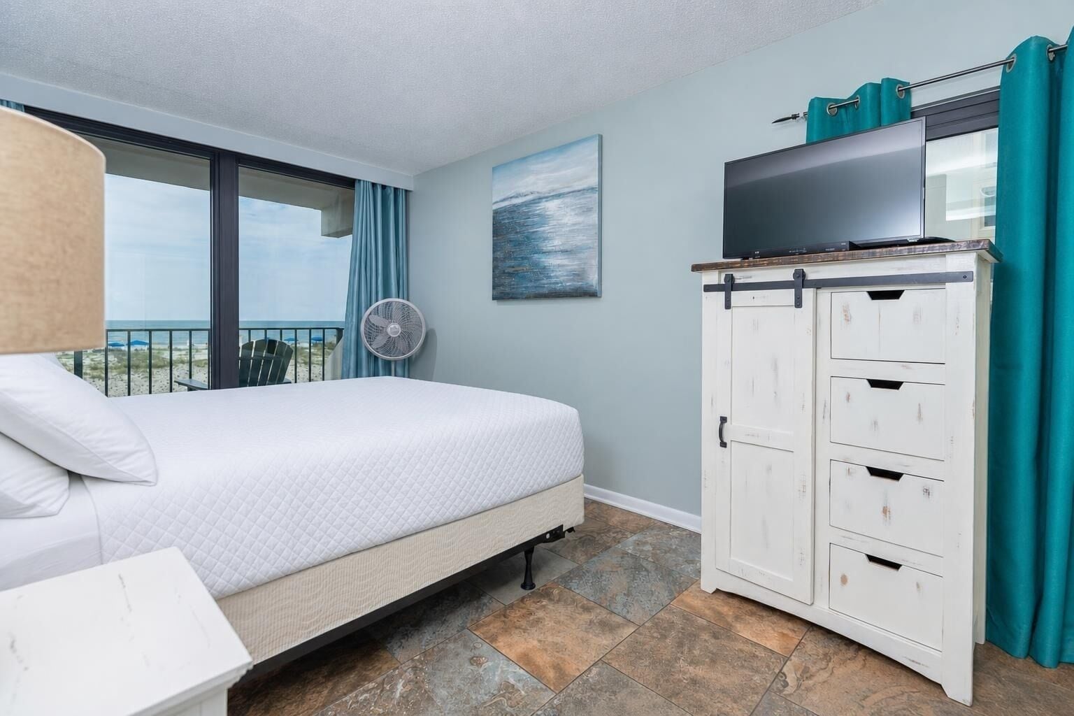 A hotel room with a bed, a TV on a cabinet, a fan, and a view of the beach.