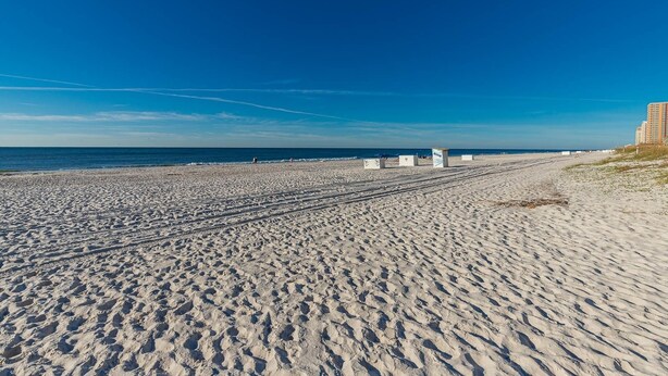 A wide sandy beach with footprints leading to the water, a clear blue sky, and a few people in the distance. - Island Winds East 109 2 Bedroom Condo (Gulf Shores)