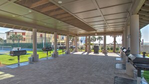 An outdoor covered patio with grilling stations, seating, and a view of a tennis court. - The Oasis 2410 Formerly Phoenix West Ii 2410 3 Bedroom Condo (Orange Beach)
