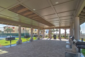 An outdoor covered patio with grilling stations, seating, and a view of a tennis court. - The Oasis 2410 Formerly Phoenix West Ii 2410 3 Bedroom Condo (Orange Beach)