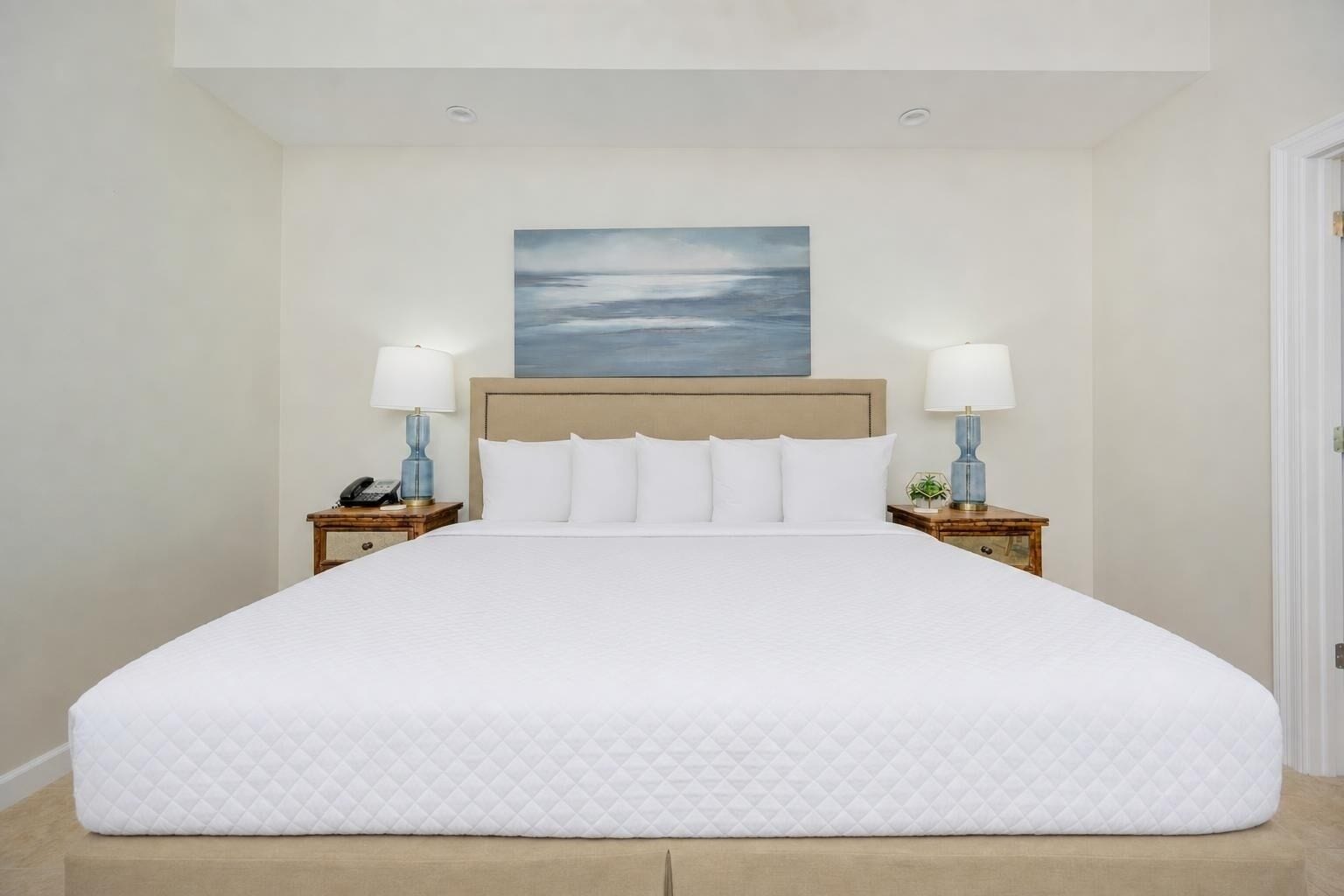 Habitación de hotel con una cama grande, dos mesitas de noche con lámparas, un cuadro en la pared y una puerta a la derecha.