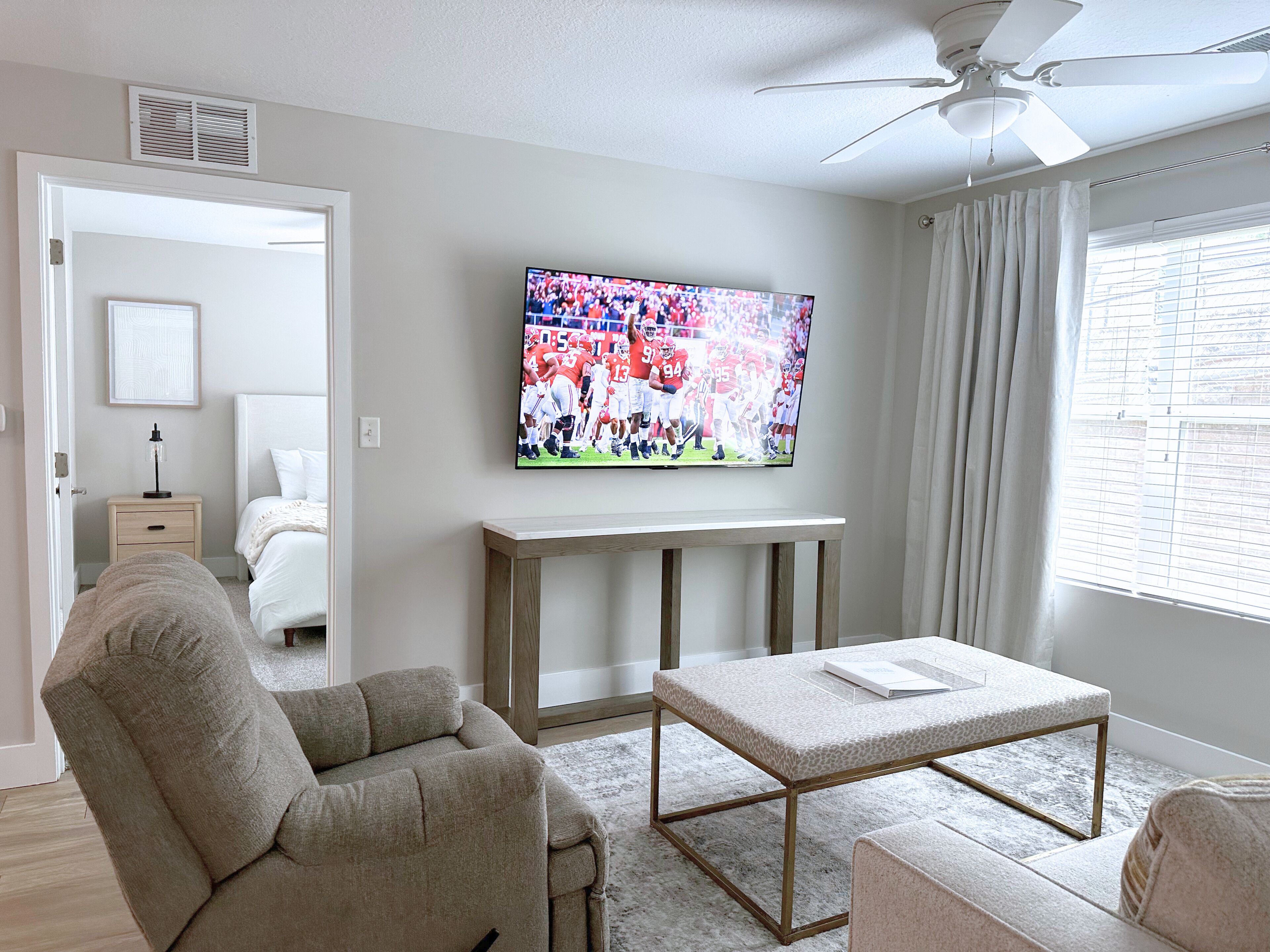 A living room with a flat-screen TV mounted on the wall, a ceiling fan, a sofa, a coffee table, and a window with blinds.