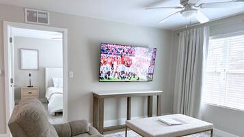 A living room with a flat-screen TV mounted on the wall, a ceiling fan, a sofa, a coffee table, and a window with blinds.