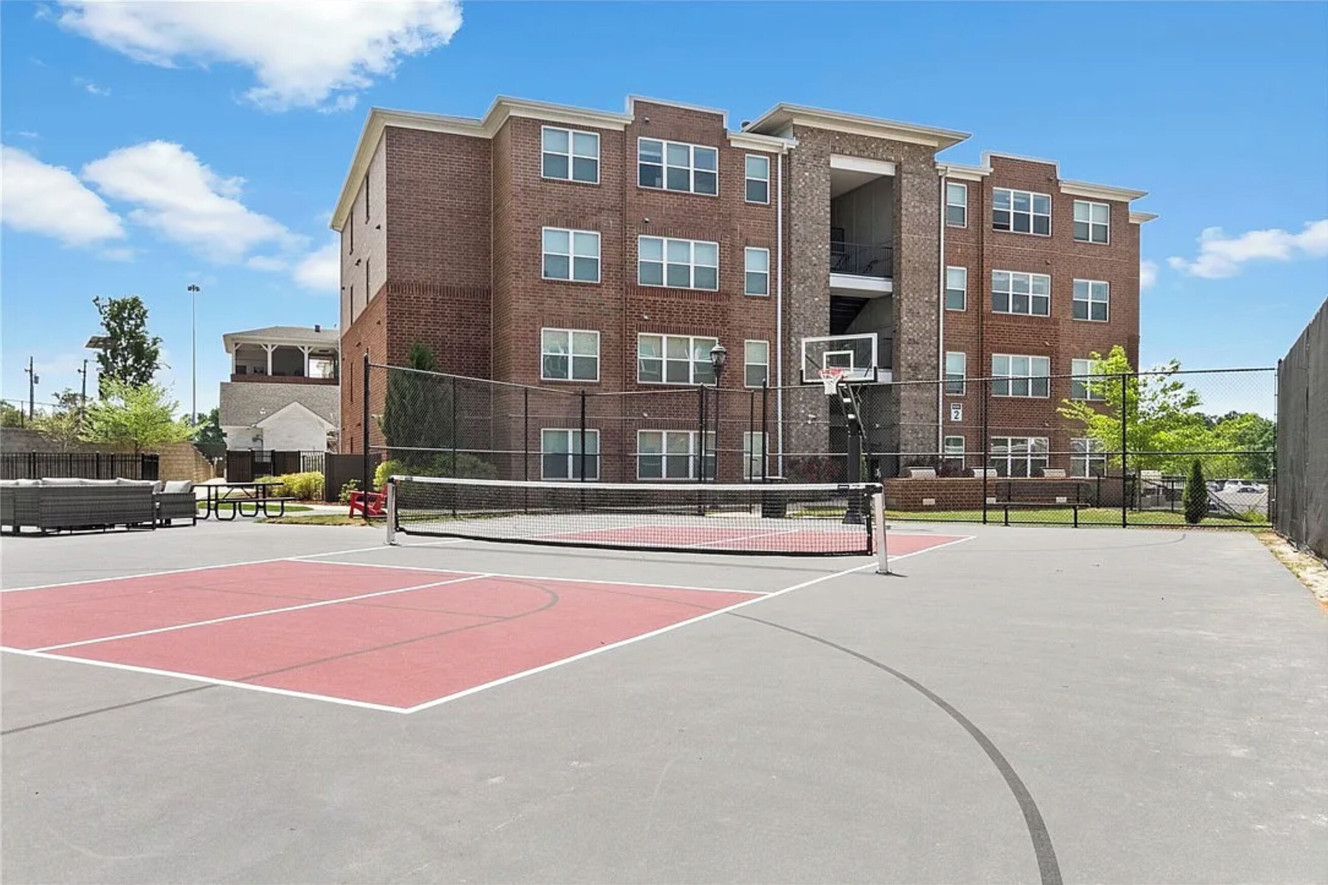 An outdoor basketball court with a net, surrounded by a fence, and a multi-story brick building in the background.