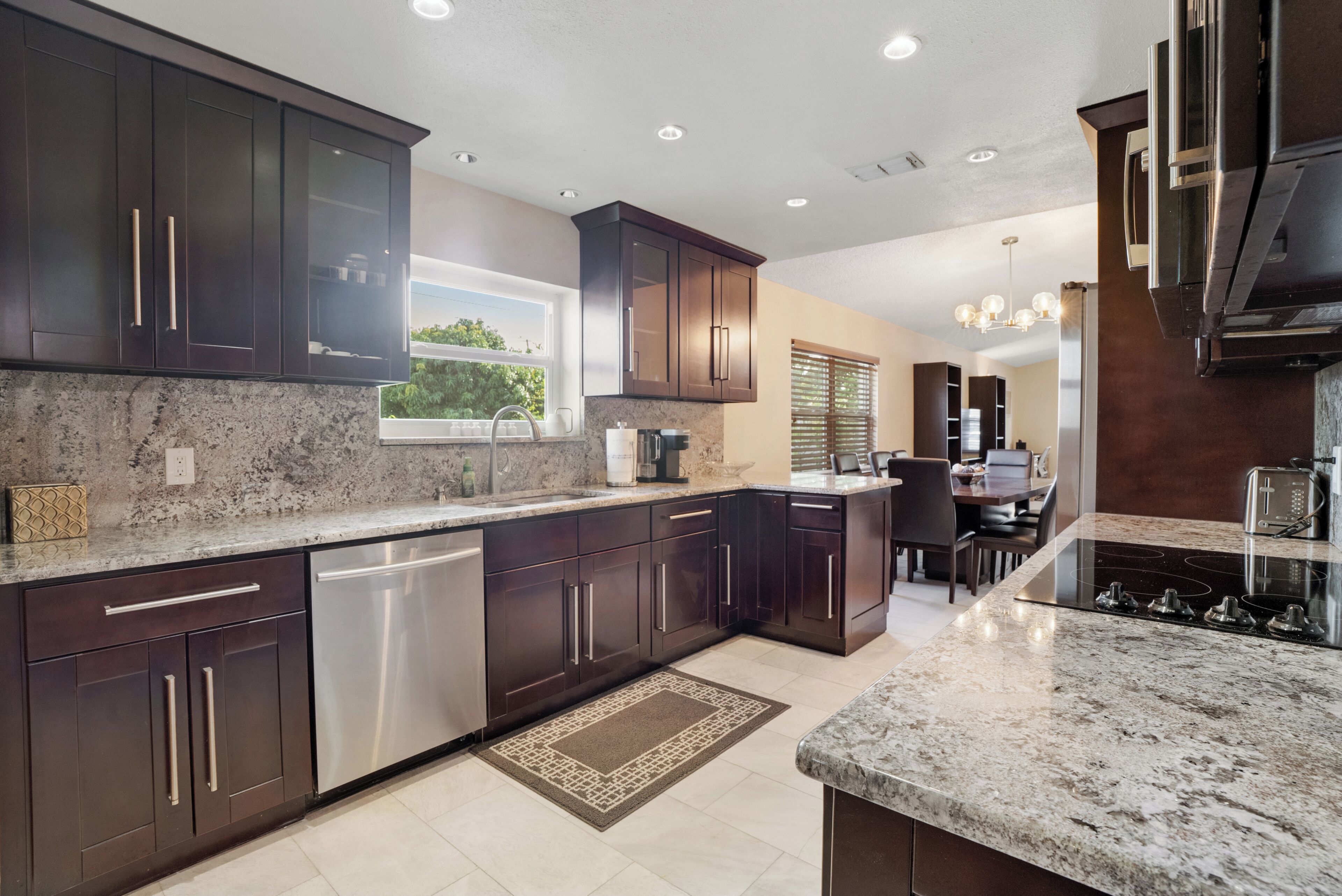 A modern kitchen with dark cabinetry, granite countertops, stainless steel appliances, and a dining area with a chandelier.