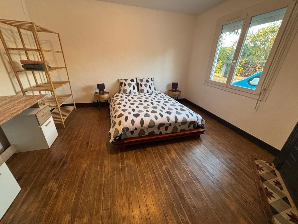 A bedroom with a wooden floor, a bed with a patterned bedspread, a bedside table, a nightstand, and a window with a view of greenery.