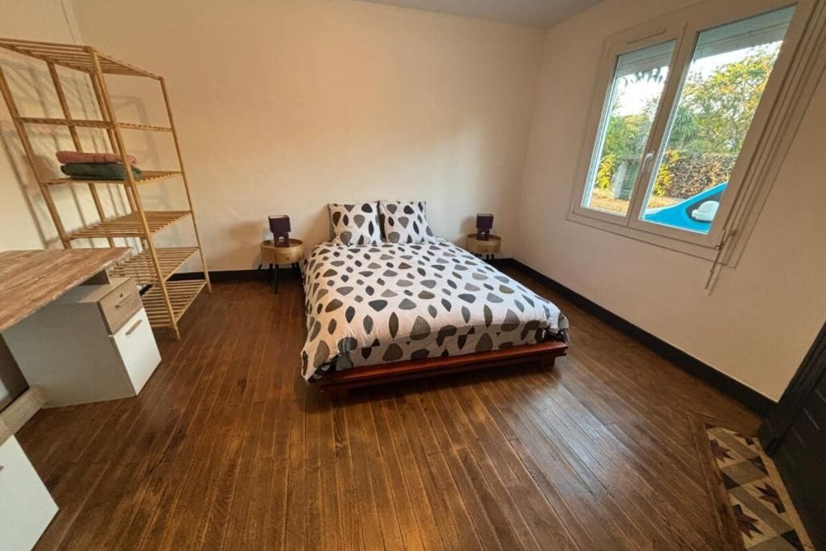 A bedroom with a wooden floor, a bed with a patterned bedspread, a bedside table, a nightstand, and a window with a view of greenery.