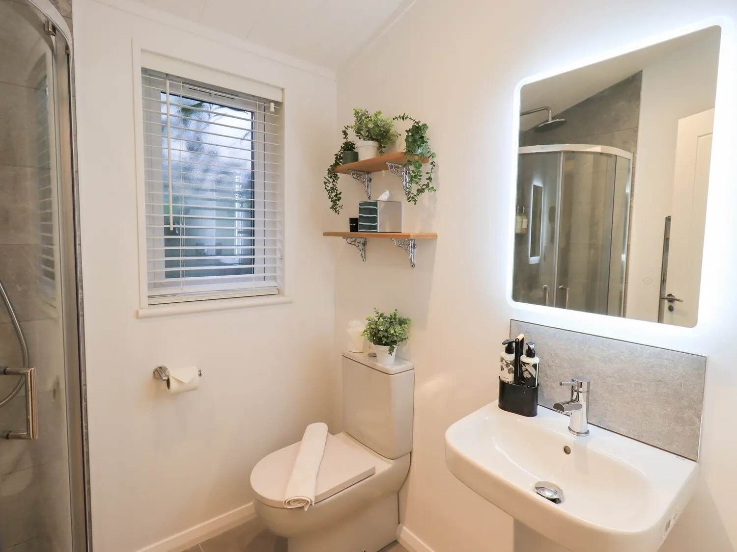 A bathroom with a toilet, sink, and shower. There are plants on a shelf above the toilet and a mirror above the sink.