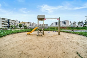 Ein Spielplatz mit Rutsche und Schaukeln, umgeben von Sand und viel Grün, mit Gebäuden im Hintergrund.