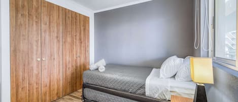 A compact bedroom with a bed, a nightstand, a window with blinds, and a wooden wardrobe.