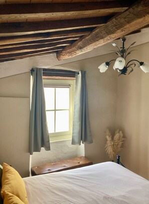 Ein Schlafzimmer mit einem Bett, einer Bank, einem Fenster mit Vorhängen, einer Pendelleuchte und einer Pflanze.