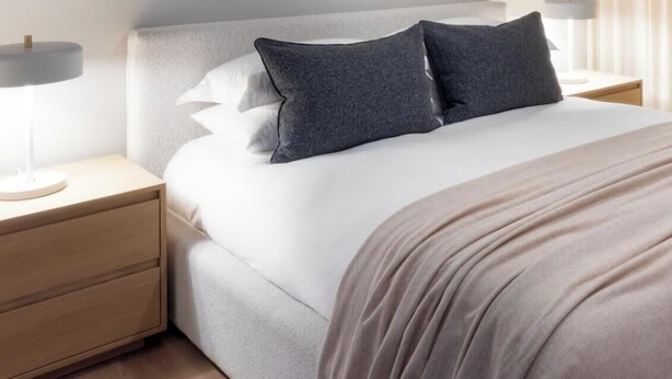 A neatly made bed with a white duvet and a beige throw blanket, flanked by two bedside tables with lamps, in a room with a large window and sheer curtains.