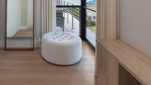 A hallway with a wooden floor, a white ottoman, and a mirror reflecting a balcony with outdoor furniture.