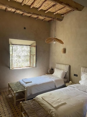 A room with two beds, a window, a hanging light fixture, and a woven bench.