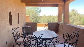 An outdoor patio with a round table and four chairs, a hanging light fixture, and a view of greenery.