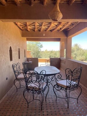 An outdoor patio with a round table and four chairs, a hanging light fixture, and a view of greenery.