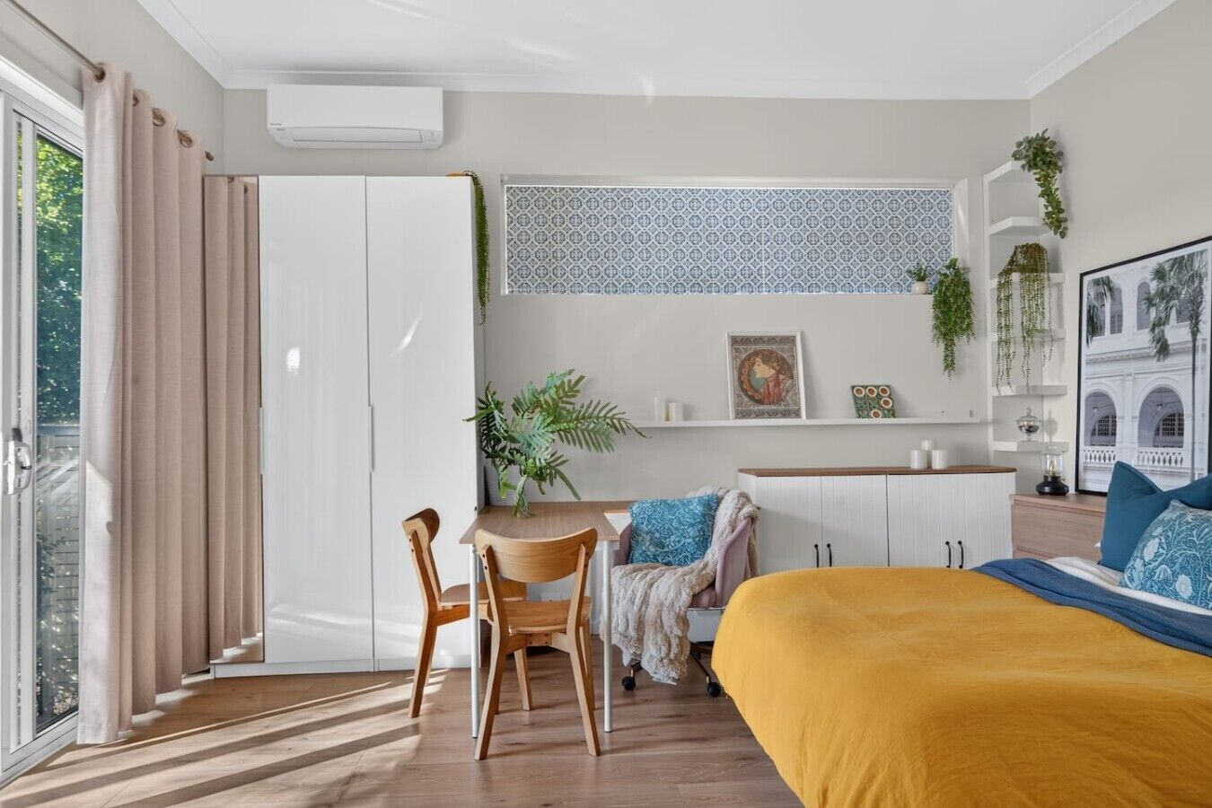 A modern bedroom with a bed, a desk, and a chair. There is a window with curtains, a plant, and a framed picture on the wall.