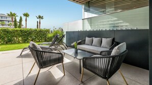 An outdoor seating area with a sofa and chairs, a glass wall, and a view of palm trees and buildings. - LOLA Beachfront Garden Apartment | Pools, Direct Beach Access, and Family-Friendly (Torremolinos)