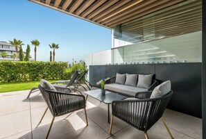 An outdoor seating area with a sofa and chairs, a glass wall, and a view of palm trees and buildings. - LOLA Beachfront Garden Apartment | Pools, Direct Beach Access, and Family-Friendly (Torremolinos)
