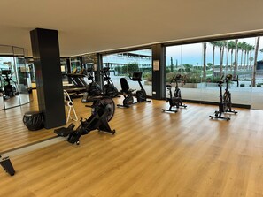 A gym with various exercise equipment, large windows, and a view of palm trees. - LOLA Beachfront Garden Apartment | Pools, Direct Beach Access, and Family-Friendly (Torremolinos)