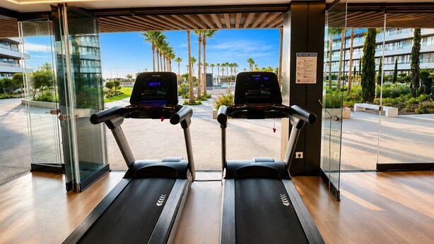 Two treadmills in a modern fitness room with glass walls, offering a view of palm trees and a clear sky. - LOLA Beachfront Garden Apartment | Pools, Direct Beach Access, and Family-Friendly (Torremolinos)