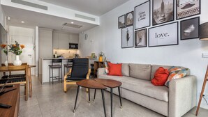 A modern living room with a sofa, coffee table, and chairs, a kitchen in the background, and a wall with framed pictures and a clock. - LOLA Beachfront Garden Apartment | Pools, Direct Beach Access, and Family-Friendly (Torremolinos)
