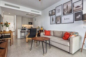 A modern living room with a sofa, coffee table, and chairs, a kitchen in the background, and a wall with framed pictures and a clock. - LOLA Beachfront Garden Apartment | Pools, Direct Beach Access, and Family-Friendly (Torremolinos)