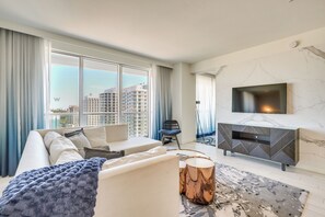 A modern living room with a large window, a sofa, a TV, and a patterned rug. - 3 High-rise Beach Condos! Views, Resort Perks (Fort Lauderdale)