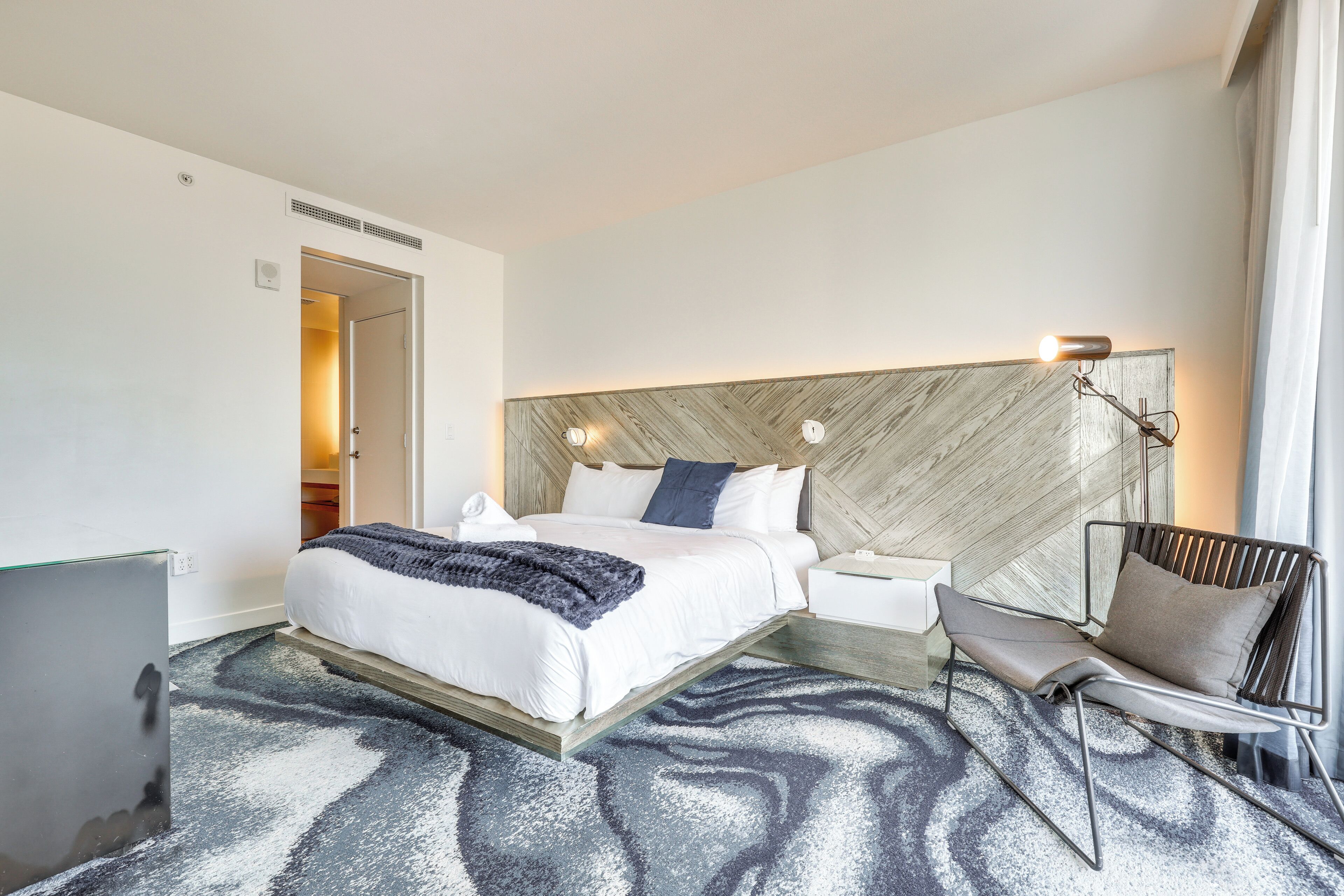 Ein modernes Hotelzimmer mit einem großen Bett, einem Schreibtisch, einem Sessel und einem Kopfteil mit integrierter Beleuchtung.