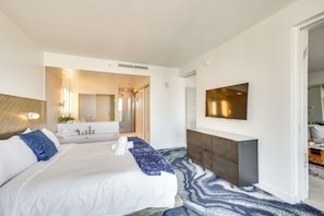 A modern hotel room with a large bed, a flat-screen TV, a wooden headboard, a built-in desk, and a bathroom with a large mirror and sink. - 3 High-rise Beach Condos! Views, Resort Perks (Fort Lauderdale)