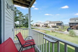 A covered porch with two red chairs, a view of a residential area with houses and a parking lot. - Saltwater Revival - Stay with Seaside Vacations! (Chincoteague Island)