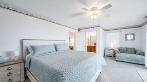 Quarto com cama, mesa de cabeceira, cômoda, ventilador de teto e porta que dá para outro espaço.